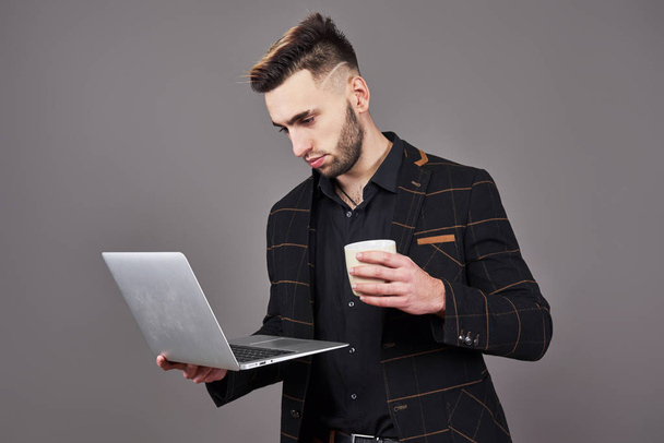 Ein bärtiger Mann in Businesskleidung unterhält sich per Smartphone und Laptop, während er vor grauem Hintergrund eine Tasse Kaffee in der Hand hält. - Foto, Bild