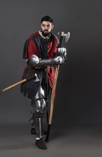Medieval knight on grey background. Portrait of brutal dirty face warrior with chain mail armour red and black clothes and battle axe. - Фото, зображення
