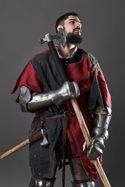 Medieval knight on grey background. Portrait of brutal dirty face warrior with chain mail armour red and black clothes and battle axe. - Φωτογραφία, εικόνα
