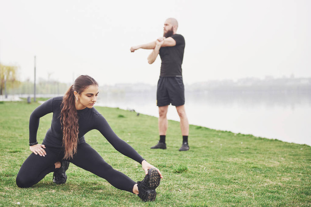 Fitness couple stretching outdoors in park near the water. Young bearded man and woman exercising together in morning. - Valokuva, kuva