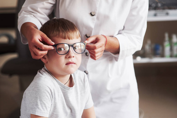 Cute kid looks into the camera while female doctor wearing new glasses to him. - Фото, зображення