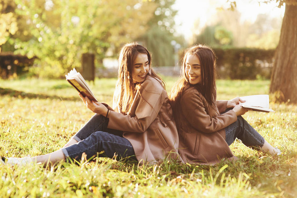Smiling brunette twin girls sitting back to back on grass and looking at each other, legs slightly bent in knees, with brown books in hands, wearing casual coat in autumn park on blurry background. - Photo, image