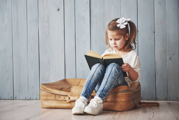 Ready to big travel. Happy little girl reading interesting book carrying a big briefcase and smiling. Travel, freedom and imagination concept. - Photo, image