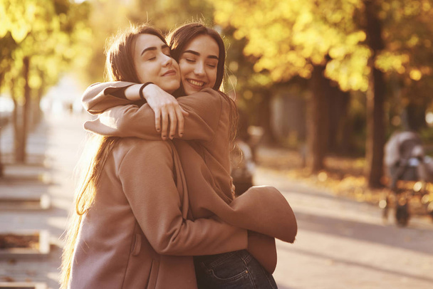 Gülümseyen güzel esmer ikiz kızların yan profili. Bulanık arka planda, sonbahar güneşli park sokaklarında birbirlerine yakın durup gündelik ceketleriyle sarılıp eğleniyorlar.. - Fotoğraf, Görsel