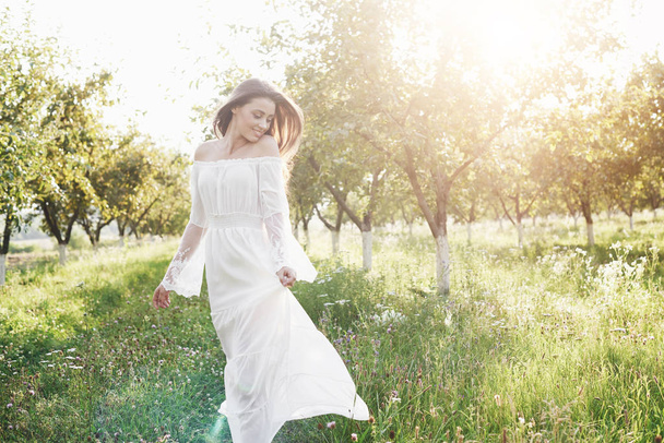 A young girl in a white long dress is walking in the garden. Beautiful sunset through the leaves of trees. - Foto, Imagem