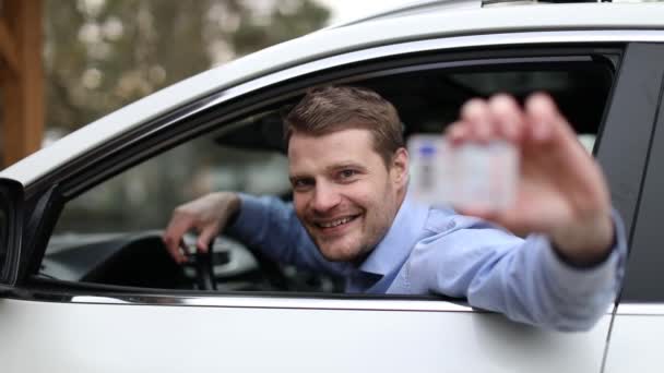 νεαρός χαρούμενος άνδρας κάθεται στο αυτοκίνητο και δείχνει το νέο δίπλωμα οδήγησης με τον αντίχειρα επάνω - Πλάνα, βίντεο