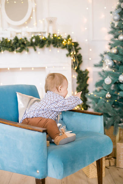 Cute little boy by the Christmas tree and fireplace decorated with garlands and gifts. Christmas mood - Foto, Imagen