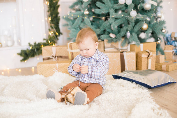 Cute little boy by the Christmas tree and fireplace decorated with garlands and gifts. Christmas mood - Фото, зображення