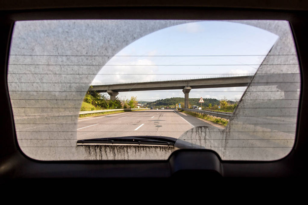 vista del coche sucio desde el interior del coche a través de la ventana trasera, ventana trasera de un coche sucio con una vista de la carretera de asfalto con marcas y un puente de carretera
. - Foto, Imagen