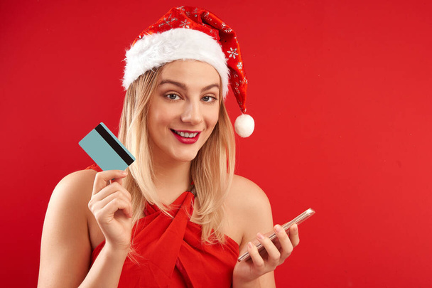 Happy excited shopping girl in santa claus christmas hat with credit card and mobile phone, buys new year gifts in online store isolated in red studio - Fotoğraf, Görsel