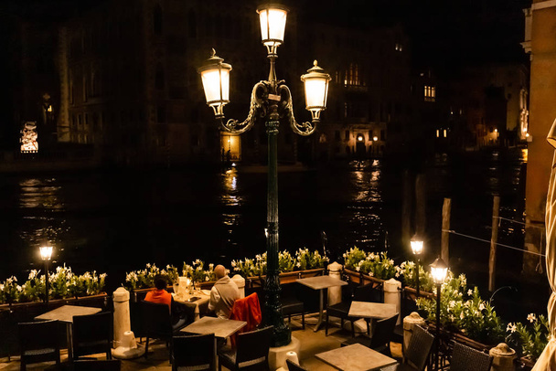 VENICE, ITALY - SEPTEMBER 24, 2019: tourists sitting at table and looking at canal at night in Venice, Italy  - Photo, image