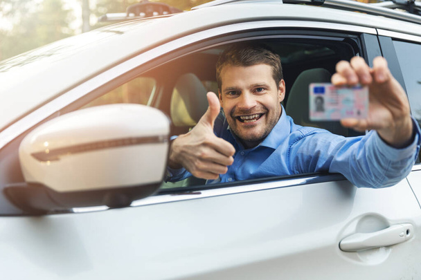 молодой улыбающийся мужчина, сидящий в машине и показывающий своего нового водителя
  - Фото, изображение