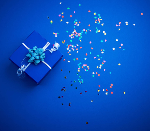 vierkante blauwe glanzende doos met een strik en veelkleurige sprankelingen op een  - Foto, afbeelding