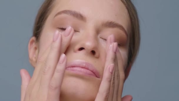 Girl looks at the camera and rubs cream on her face close up - Imágenes, Vídeo