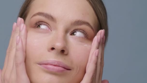 Girl looks at the camera and rubs cream on her face close up - Imágenes, Vídeo