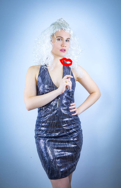 Close up portrait cheerful comic attractive beautiful girl in a wig wearing silver dress and having fun using fake lips on a wooden stick, isolated on a light background - Fotó, kép