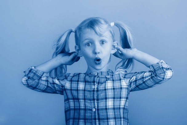 Funny Caucasian young girl making faces in front of camera. Child holding pulling dragging ears. Kid expressing emotions. April fool day concept. Toned with classic blue 2020 color. - Photo, Image
