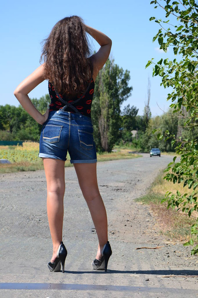 hátsó lövés vonzó barna hajú fiatal nő viselt kék farmer rövidnadrág fekete top cipőben magas sarkú cipő áll a vidéki úton, és vár egy autó - Fotó, kép