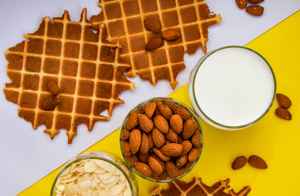 homemade waffles with almond nuts in the glass and almond chips and almond milk in the glass on the white and yellow background, copy space, top view, Making almond milk from dried almonds - Photo, Image
