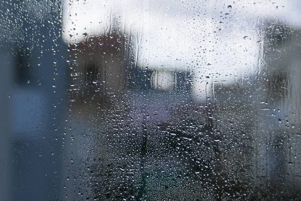 Nedves ablaküveg vízcseppekkel és homályos házalakzatokkal a sápadt ég előtt - Fotó, kép