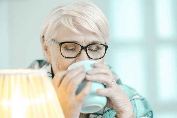 Senior Woman Wrapped In A Checkered Plaid Is Drinking Tea At Home - Foto, Imagem