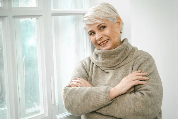 Grey-haired Senior Woman Standing At The Window And Looking Asid - Foto, Imagen