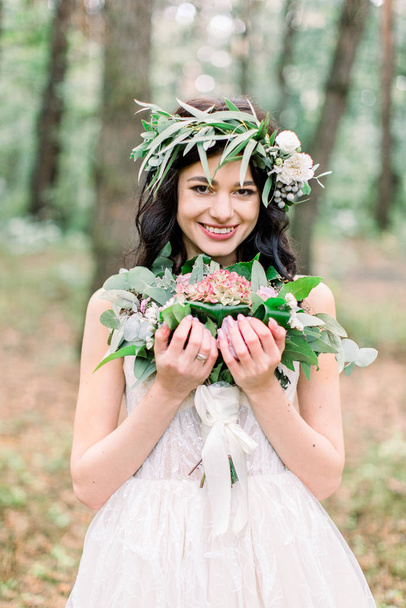 Wedding ceremony in rustic style. Beautiful smiling bride with green wreath on the head, with bouquet in her hand, posing in pine forest - Foto, immagini