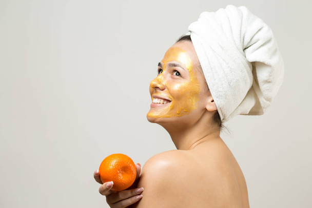 Piękny portret kobiety w białym ręczniku na głowie ze złotą maską odżywczą na twarzy. Oczyszczanie skóry ekologiczny kosmetyczny spa zrelaksować koncepcję. Dziewczyna stoi z tyłu trzymając pomarańczowy mandarynkę. - Zdjęcie, obraz