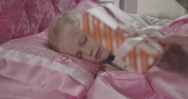 Close-up portrait of pretty little Caucasian girl sleeping in pink bed as male hand putting Christmas present on pillow. Holidays season, miracle, happiness, childhood. Cinema 4k ProRes HQ. - Felvétel, videó
