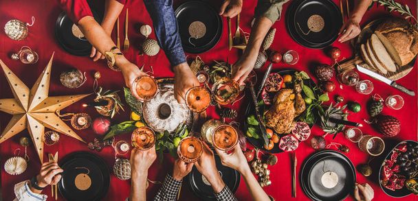 Friends celebrating Christmas. Flat-lay of people clinking glasses with rose wine over festive table with red cloth with roasted chicken, bundt cake, fruit, decorations, top view. Winter holiday party - Fotoğraf, Görsel
