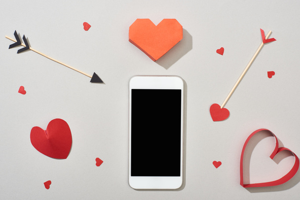 Top view of smartphone with blank screen and heart shaped papers with arrows on grey background - Фото, изображение