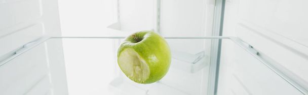 Panoramic shot of bitten off green apple on shelf of refrigerator with open door isolated on white - Photo, Image