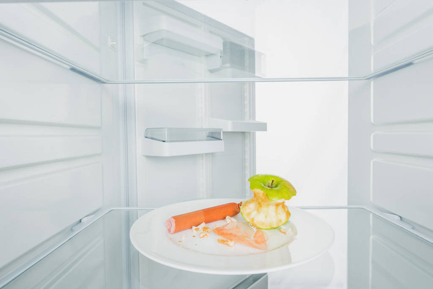 Pomme grignotée aux saucisses et miettes dans une assiette au réfrigérateur avec porte ouverte isolée sur fond blanc
 - Photo, image