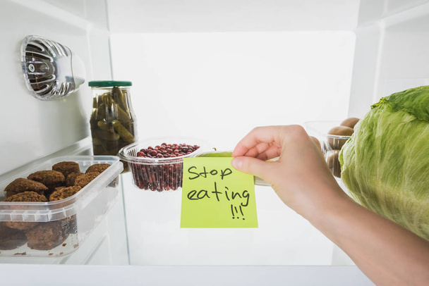 Cropped view of woman holding card with stop eating lettering with food in fridge at background, isolated on white - Photo, Image