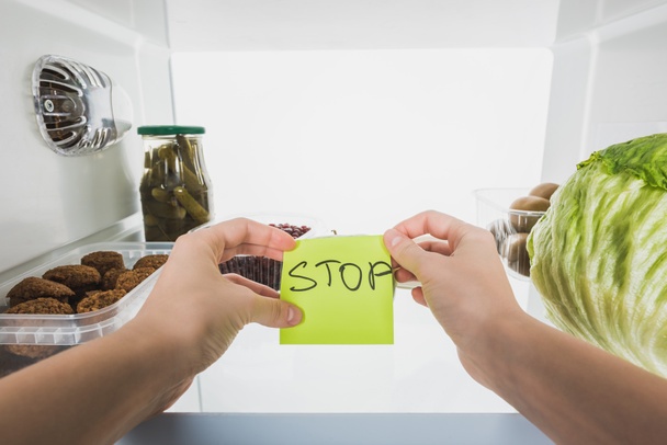 Ausgeschnittene Ansicht einer Frau mit Karte mit Stop-Schriftzug mit Lebensmitteln im Kühlschrank im Hintergrund, isoliert auf weiß - Foto, Bild