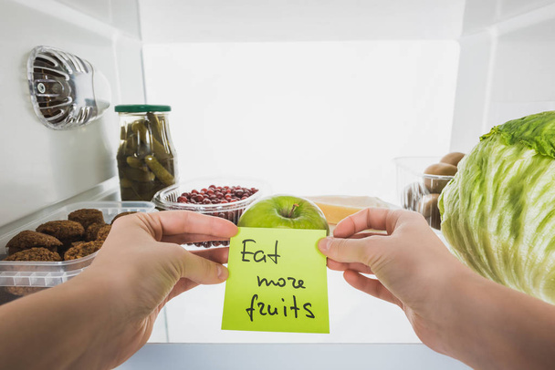 Vista recortada de la mujer que sostiene la tarjeta con comer más letras de frutas con alimentos en la nevera en el fondo, aislado en blanco
 - Foto, Imagen