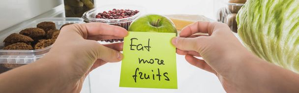 Panoramic shot of woman holding card with eat more fruits lettering with food in fridge at background, isolated on white - Photo, Image