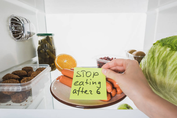 Vista recortada de la mujer con dejar de comer después de seis letras en la tarjeta con alimentos en nevera aislado en blanco
 - Foto, Imagen