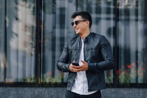 young stylish guy in glasses in black leather jacket with phone on glass background - Фото, зображення