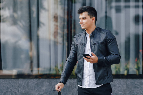 young stylish guy in glasses in black leather jacket with phone on glass background - Foto, Imagen