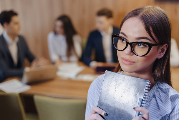młody atrakcyjny stylowy pracownik biurowy w okularach z notatnikiem w rękach na tle pracujących kolegów - Zdjęcie, obraz