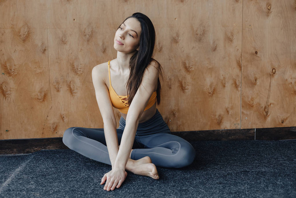 Young attractive fitness girl sitting on the floor near the window on the background of a wooden wall, resting on yoga classes - Fotografie, Obrázek