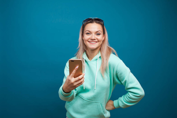 Cheerful blonde with a mobile phone. Portrait of a young positive woman in a blue hoodie - Fotografie, Obrázek