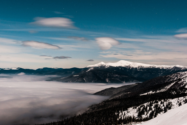 живописный вид снежных гор с соснами и белыми пушистыми облаками в темном небе вечером
 - Фото, изображение
