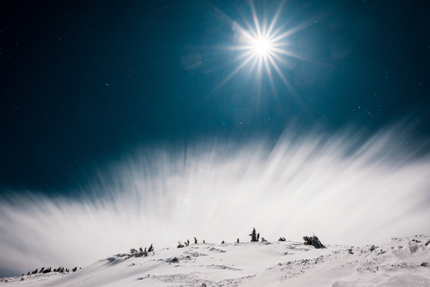 γραφική θέα του βουνού που καλύπτεται με χιόνι και πεύκα κατά σκοτεινό ουρανό με λευκό σύννεφο και ήλιο - Φωτογραφία, εικόνα