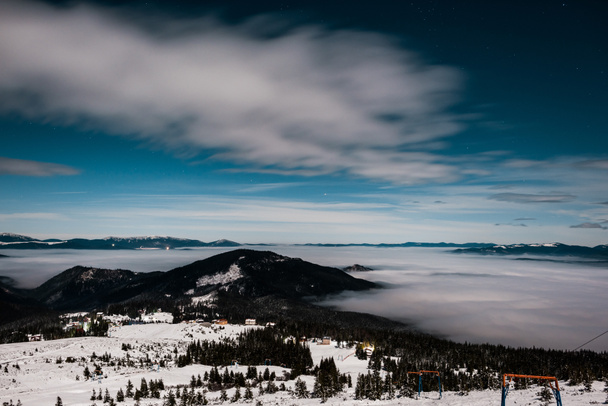 luonnonkaunis näkymä lumiset vuoret mäntyjä ja valkoinen pörröinen pilvet pimeässä taivaalla illalla
 - Valokuva, kuva