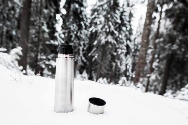 Vakuumflasche mit Heißgetränk auf schneebedecktem Hügel in der Nähe von Kiefernwald - Foto, Bild
