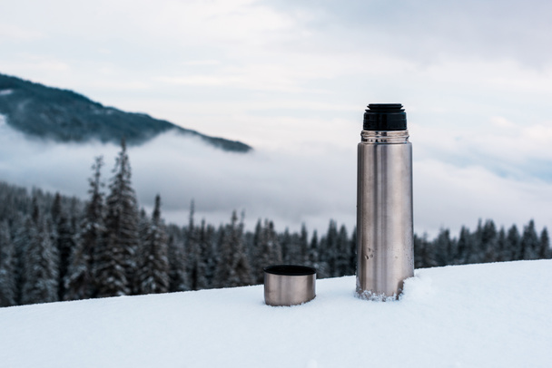 open metallic vacuum flask in snowy mountains with pine trees and white fluffy clouds - Photo, Image