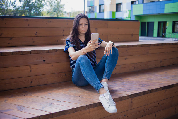 A brunette girl, a student with a large wrist watch, sits on wooden steps with legs and thoughtfully checks smartphone. - Фото, зображення