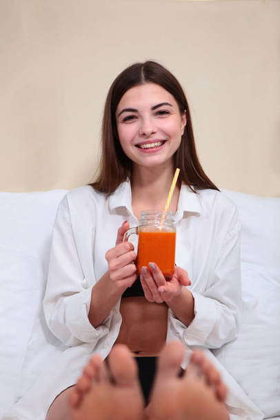 Beautiful girl drinking juice in bed and smiling. Breakfast concept at home or in the hotel. Copy space. Legs out of focus. - Foto, Imagem